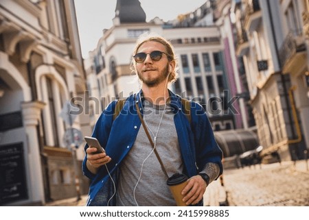 Young beard handsome man drinking coffee and listening music in headphones while walking on the city streets, exploring the old town, finding the direction route on application online