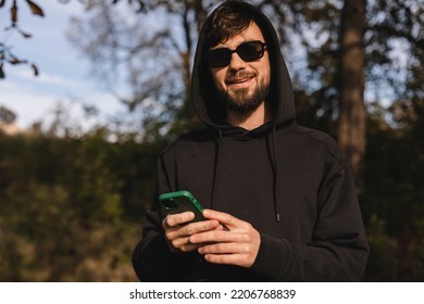 Young Beard Man In Black Hoody And Glasses Use Mobile Cell Phone Chat Rest Relax In City Park Sunshine Lawn Outdoors On Nature. Urban Lifestyle Leisure Concept. Man Look At Camera.