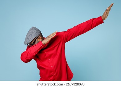 Young bearded african american man 20s in red shirt eyeglasses cap doing dab hip hop dance hands gesture, youth sign hiding and covering face isolated on pastel blue color background studio portrait
