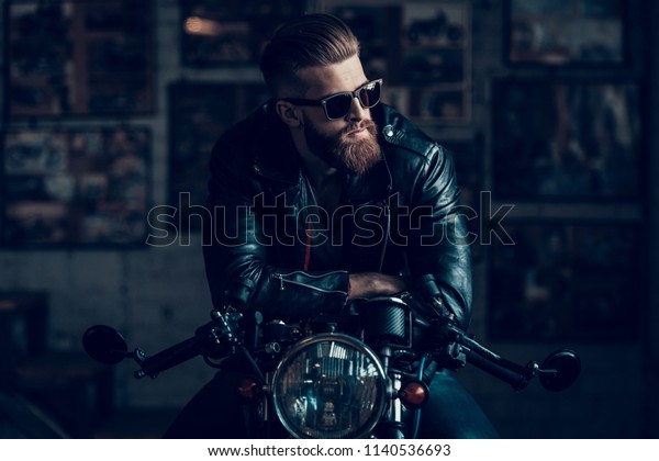 Young\
Bearded Biker Sitting on Motorcycle in Garage. Indoor Garage. Young\
Mechanic in Garage. Parts of Motorcycle. Man in Checkered Shirt.\
Man on Vintage Bike. Biker Lifestyle\
Concept.