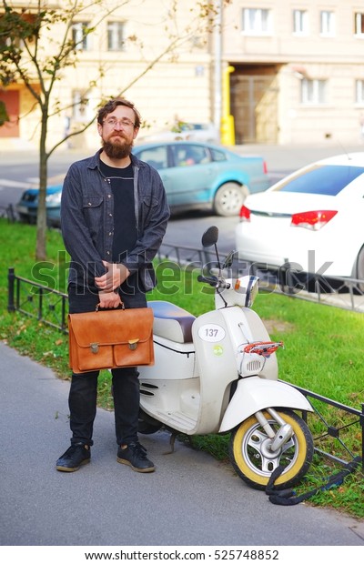 Young bearded business man in\
glasses with a briefcase standing near a motor scooter in his\
yard