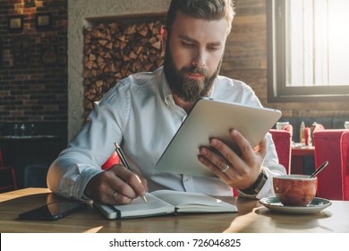 Young bearded businessman sits in cafe, home at table, looking at screen of tablet computer and writes in notebook.Man is working, studying.Online education,marketing, training. E-learning,e-commerce. - Powered by Shutterstock