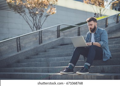 Young bearded businessman sits on steps, using laptop and looks on his screen.Hipster man is working, blogging, chatting online,checking email. Student learning online. Summer sunny day. Lifestyle. - Powered by Shutterstock