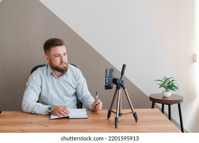 Young bearded businessman working in office sitting at table using smartphone for an online meeting making notes in diary. Freelancer works remotely. Student is preparing for seminar. Online marketing