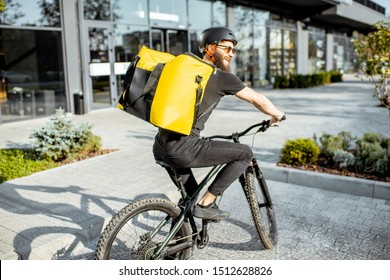 Young bearded courier delivering food with a yellow thermal backpack, riding a bicycle in the city. Food delivery service concept
