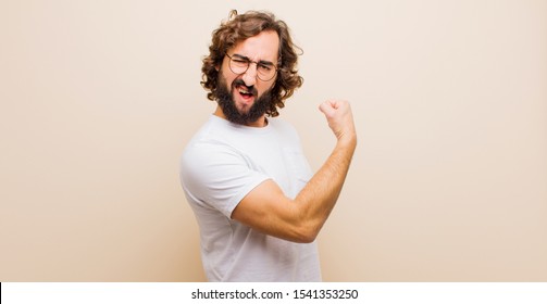 young bearded crazy man feeling happy, satisfied and powerful, flexing fit and muscular biceps, looking strong after the gym against flat color wall