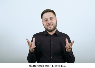 Young Bearded Dark Haired Man In Black Stylish Shirt On White Background, Happy Male Holding His Hands Wide As If Explaining Something. The Concept Of Speakers, People Who Prove An Opinion