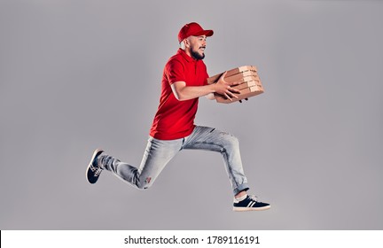 Young bearded delivery man in a red uniform runs in a jump flight holds boxes with pizza isolated on gray background. Fast home delivery of pizza.