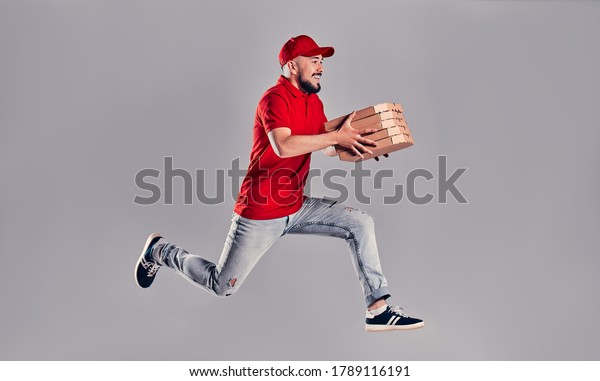 Young bearded delivery man in a red uniform runs\
in a jump flight holds boxes with pizza isolated on gray\
background. Fast home delivery of\
pizza.