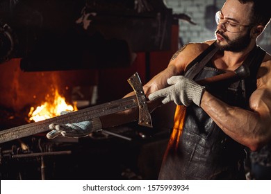 young bearded forger man studying handmade metal in workshop near furnace, wearing brown leather apron, gloves and protective eyeglasses - Powered by Shutterstock