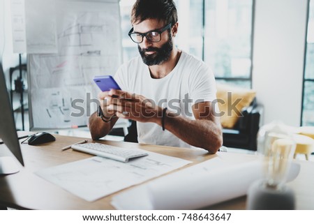 Young bearded graphic designer wearing eye glasses and working at modern loft studio-office.Man using smartphone.Blurred background. Horizontal