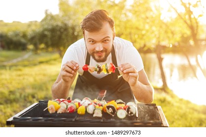 Young bearded guy in apron smiling and looking at camera while cooking tasty shish kebab on grill in countryside - Powered by Shutterstock