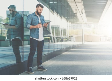 Young bearded hipster businessman stands on city street, holds cup of coffee and uses tablet computer.In background is modern glass building. Man working, blogging, chatting online.Social media. - Powered by Shutterstock