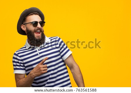 Young bearded hipster in cap and sunglasses pointing happily away on orange background. 