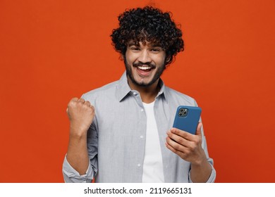 Young Bearded Indian Man 20s Years Old Wears Blue Shirt Hold In Hand Use Mobile Cell Phone Doing Winner Gesture Celebrate Clenching Fists Say Yes Isolated On Plain Orange Background Studio Portrait