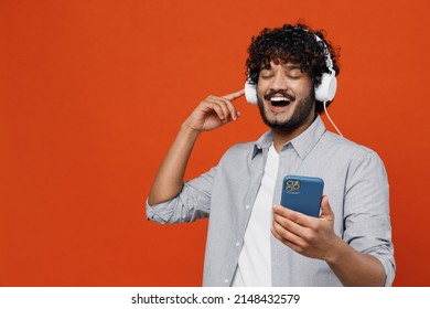 Young Bearded Indian Man 20s Years Old Wears Blue Shirt Listen Music In Headphones Dance Sing Song Have Fun Enjoy Hold In Hand Use Mobile Cell Phone Isolated On Plain Orange Background Studio Portrait