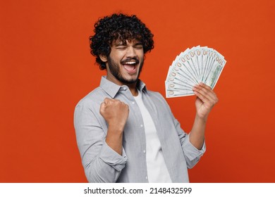 Young bearded Indian man 20s years old wears blue shirt holding fan of cash money in dollar banknotes doing winner gesture celebrate clenching fists isolated on plain orange background studio portrait