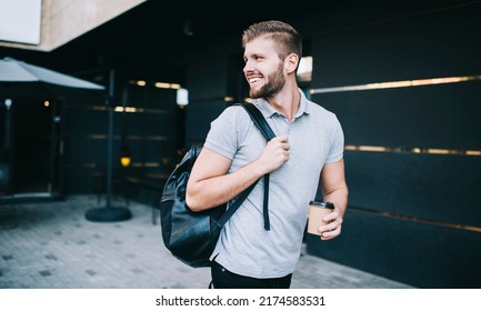 Young bearded male in casual clothes with leather backpack standing near building with paper cup of coffee and looking away - Powered by Shutterstock