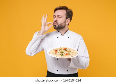 Young bearded male chef cook or baker man in white uniform shirt isolated on yellow background. Cooking food concept. Mock up copy space. Hold italian pizza on plate making okay taste delight sign