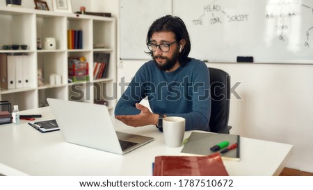 Young bearded male teacher sitting at his workplace and teaching Computer Science online. Looking at webcam and talking with audience. Stay home. Focus on man. E-learning. Distance education