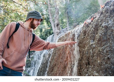 Young Bearded Man With A Bucket Hat Touching The Water That Runs Through A Wall Of A Waterfall