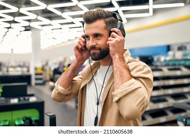 Young Bearded Man Buying Headphones While Listening To Music In A Home Appliances, Electronics And Gadgets Store.