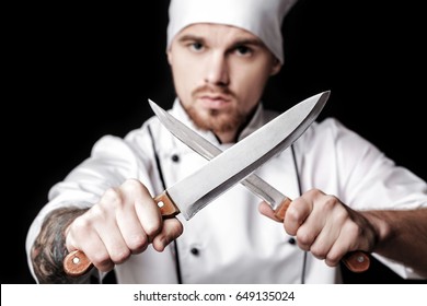 Young bearded man chef In white uniform holds Two knives on  black background. Focus on knives