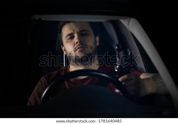 A young bearded man drives a car holding a\
bottle of beer in his hand. Drunk driving on highways at night.\
Dangerous driving, breaking the law. Drinking alcohol while\
driving. Drunk driver\
concept.