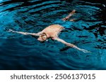 A young bearded man floats calmly on the surface of a dark blue body of water, with his arms outstretched and his eyes closed. 