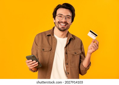 Young bearded man in glasses holds credit card and smartphone isolated on orange background. Online shopping. Online payment. - Shutterstock ID 2027914103