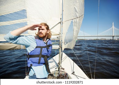 Young Bearded Man In Lifejacket Floating On Yacht And Looking Forwards With His Hand By Forehead