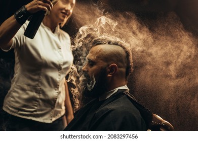 Young bearded man with a mohawk getting haircut by hairdresser while sitting in chair at barbershop and smoking a cigarette - Powered by Shutterstock