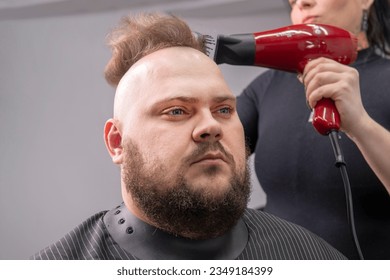 Young bearded man with a mohawk getting haircut by hairdresser while sitting in chair at barbershop. A hairdresser girl makes a mohawk to a guy.