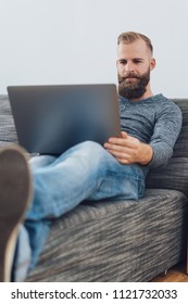 Young Bearded Man Relaxing With His Feet Up On A Comfortable Couch At Home As He Works On His Laptop Computer