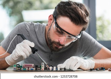 Young Bearded Man Repairing Motherboard from PC. Repair Shop. Worker with Tools. Computer Hardware. Magnifying Glass. Soldering Iron. Digital Device. Laptop on Desk. Electronic Devices Concept. - Powered by Shutterstock