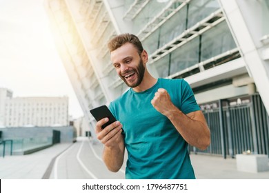 Young Bearded Man Showing Sincere Excitement About His Favorite Team Victory. Guy Being Happy Winning A Bet In Online Sport Gambling Application On His Mobile Phone. 