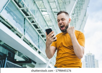 Young Bearded Man Showing Sincere Excitement About His Favorite Team Victory. Guy Being Happy Winning A Bet In Online Sport Gambling Application On His Mobile Phone. Stadium On The Background.