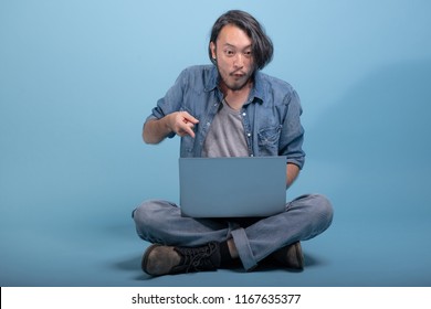 Young Bearded Man Sit Down On Floor Using Computer In Blue Background. Excited Asian Young Hipster Using Laptop, Full Body Shot. Young Generation Hipster Working Concept.