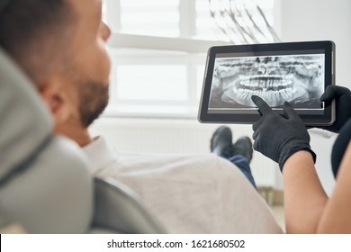 Young bearded man sitting in dental chair and looking at teeth picture on tablet that holding dentist in black gloves. Professional doctor examining oral cavity of male patient. - Powered by Shutterstock