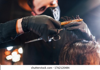 Young Bearded Man Sitting And Getting Haircut In Barber Shop By Guy In Black Protective Mask.