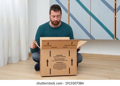 Young bearded man sitting on wooden floor of his casual apartment and looking inside a carton box with a disappointed gesture due to a wrong delivery from an online store.