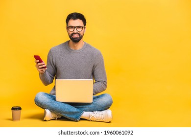 Young Bearded Man Sitting On The Floor With Laptop And Talking At Phone. Isolated Over Yellow Background.