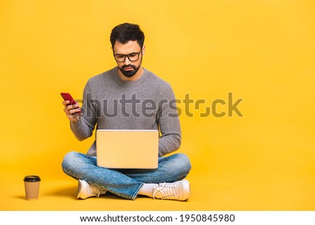 Young bearded man sitting on the floor with laptop and talking at phone. Isolated over yellow background.