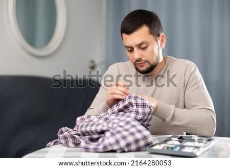 Young bearded man sitting at table in living room and sewing on buttons on sleeve of his shirt ..