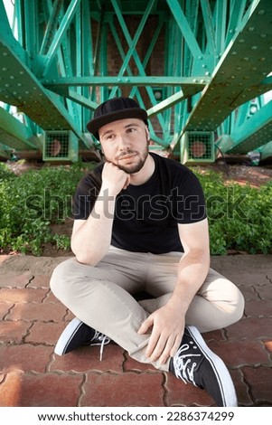 Young bearded man sitting and thinking on a pavement under a green bridge. One guy dressed casually relaxing on a street sidewalk