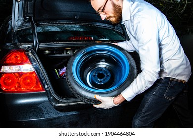 A Young Bearded Man Taking A Spare Wheel Out Of A Boot, Of A Trunk Of A Black Car. Changing A Car Tyre, Tire At Night.