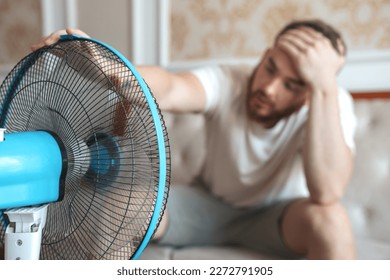 Young bearded man using electric fan at home, sitting on couch cooling off during hot weather, suffering from heat, high temperature - Shutterstock ID 2272791905