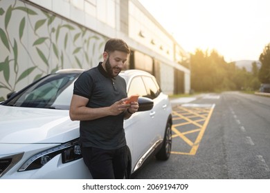 Young bearded man using phone supported on white parked car. He is wearing black clothes. He is smiling. He is using orange smartphone. He is near industrial building during sunset. - Powered by Shutterstock