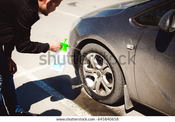 young bearded man washes his car\'s\
wheel rims, spraying water from spray, in street\
parking