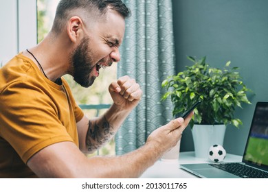 Young Bearded Man Watching Soccer Play Online On The Laptop And Celebrating Favorite Team Victory. Guy Being Happy Winning A Bet In Online Sport Gambling Application On Mobile Phone. 